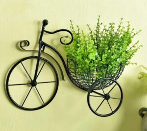 Black vintage bicycle wall mounted planter made of iron with a green leafy plant in the wire basket on a light yellow wall.